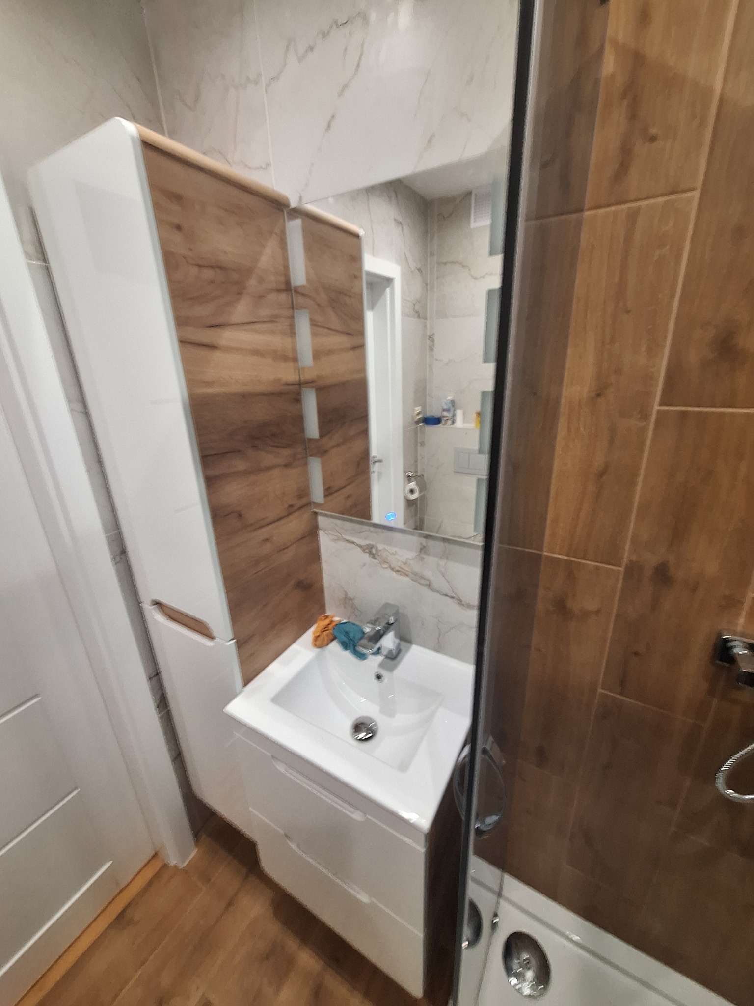 Nowoczesna łazienka z zabudową umywalkową w bieli i drewnie, widok z kabiny prysznicowej na umywalkę, szafkę i lustro, z detalami płytek imitujących marmur.