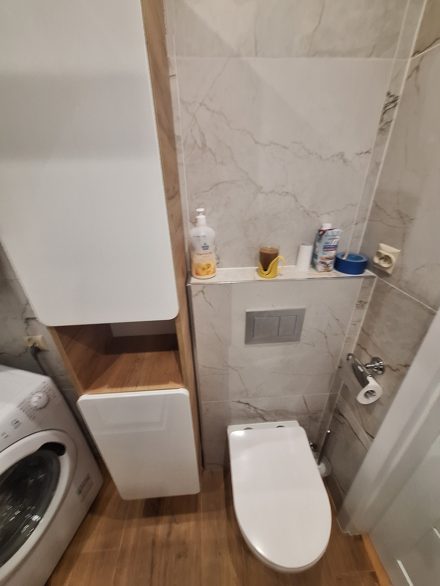 Nowoczesna toaleta podwieszana z białym siedziskiem, szara płytka imitująca marmur na ścianach, obok pralka i szafka łazienkowa z drewnianymi elementami, na półce nad toaletą różnorodne akcesoria...