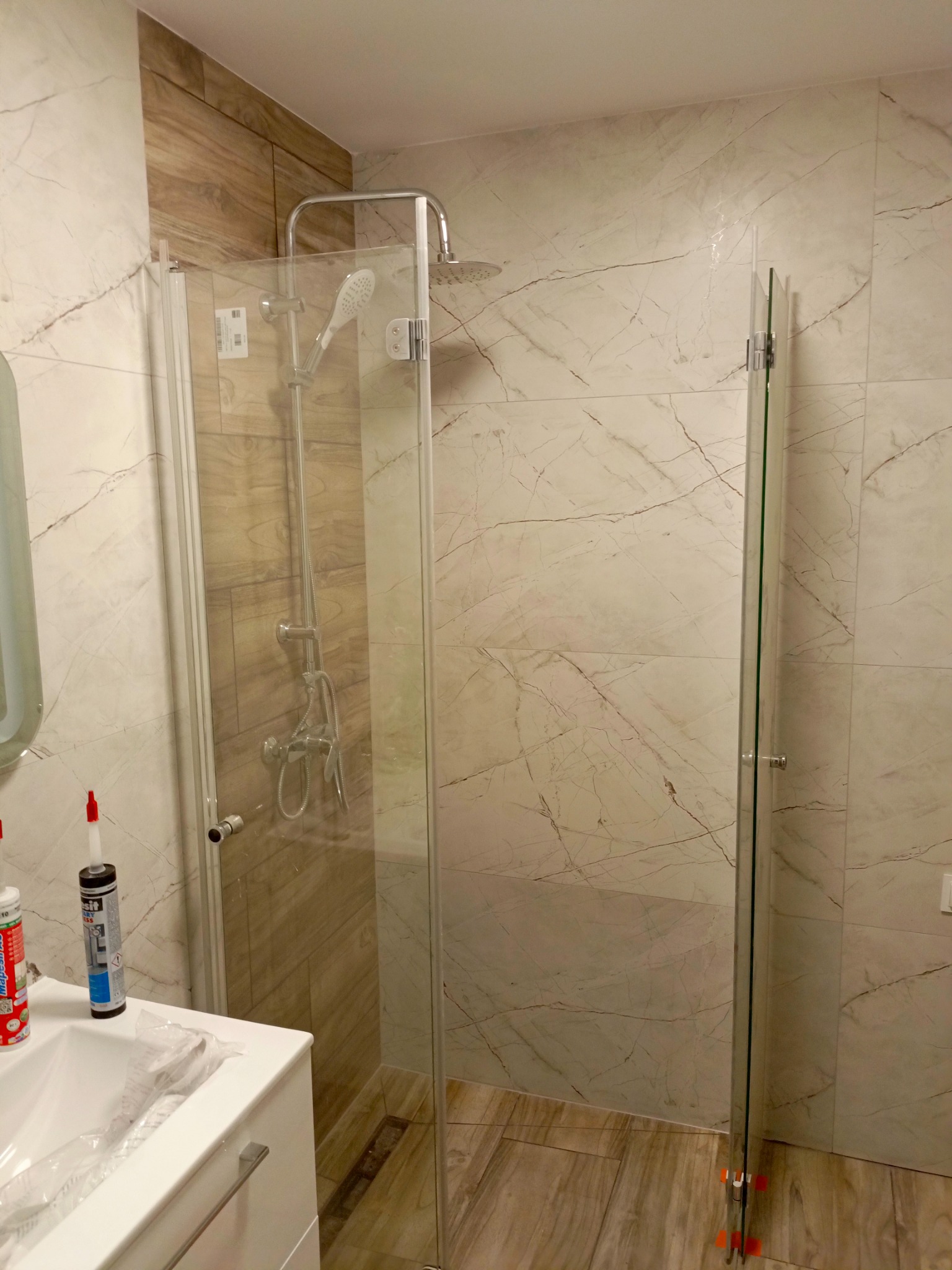 Nowoczesna kabina prysznicowa z chromowaną armaturą i deszczownicą, zamontowana w łazience wykończonej płytkami imitującymi marmur i drewno, widoczne silikony sanitarne na umywalce.