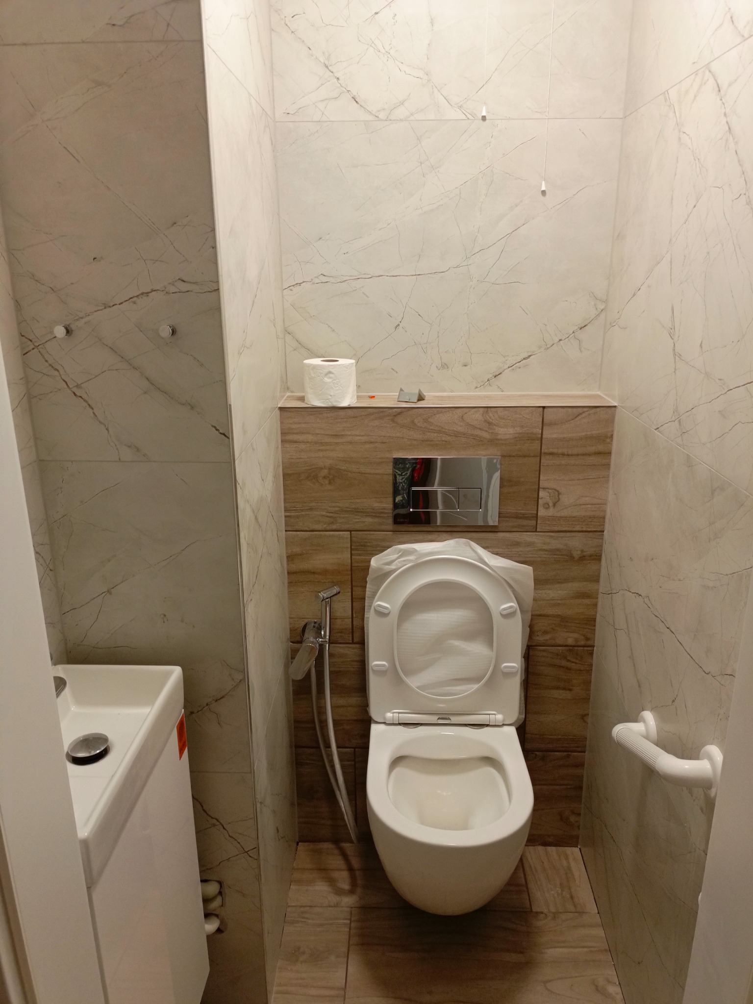 Nowoczesna toaleta w trakcie montażu, z częściowo zdjętą folią ochronną na desce sedesowej, marmurowe płytki na ścianach i drewnopodobne na podłodze, widoczny bidetta i umywalka.