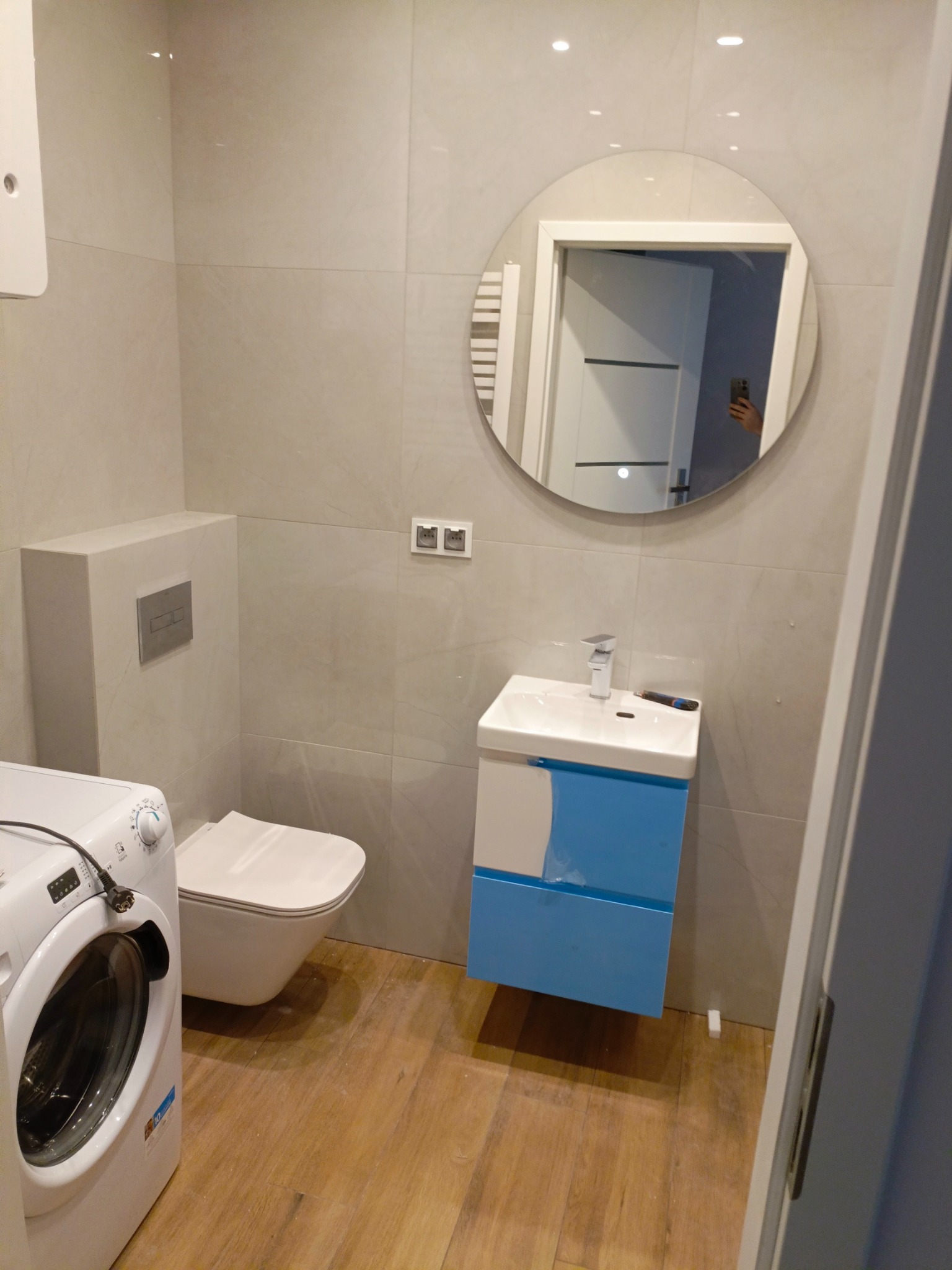 Nowoczesna łazienka z beżowymi płytkami, wiszącą toaletą, szafką umywalkową w kolorze błękitnym, okrągłym lustrem i pralką Candy z widocznym wtykiem.