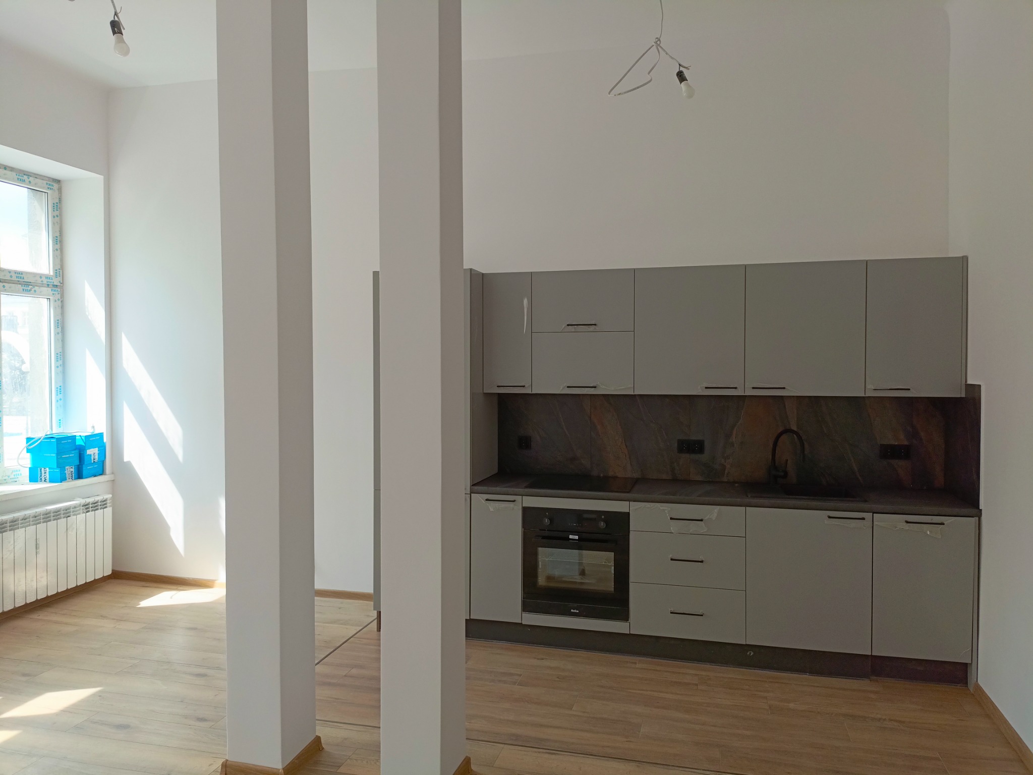 Nowoczesna szara kuchnia z piekarnikiem Amica i czarnym blatem, widok z perspektywy pokoju z trzema białymi filarami, okno z niebieskimi pojemnikami w tle.