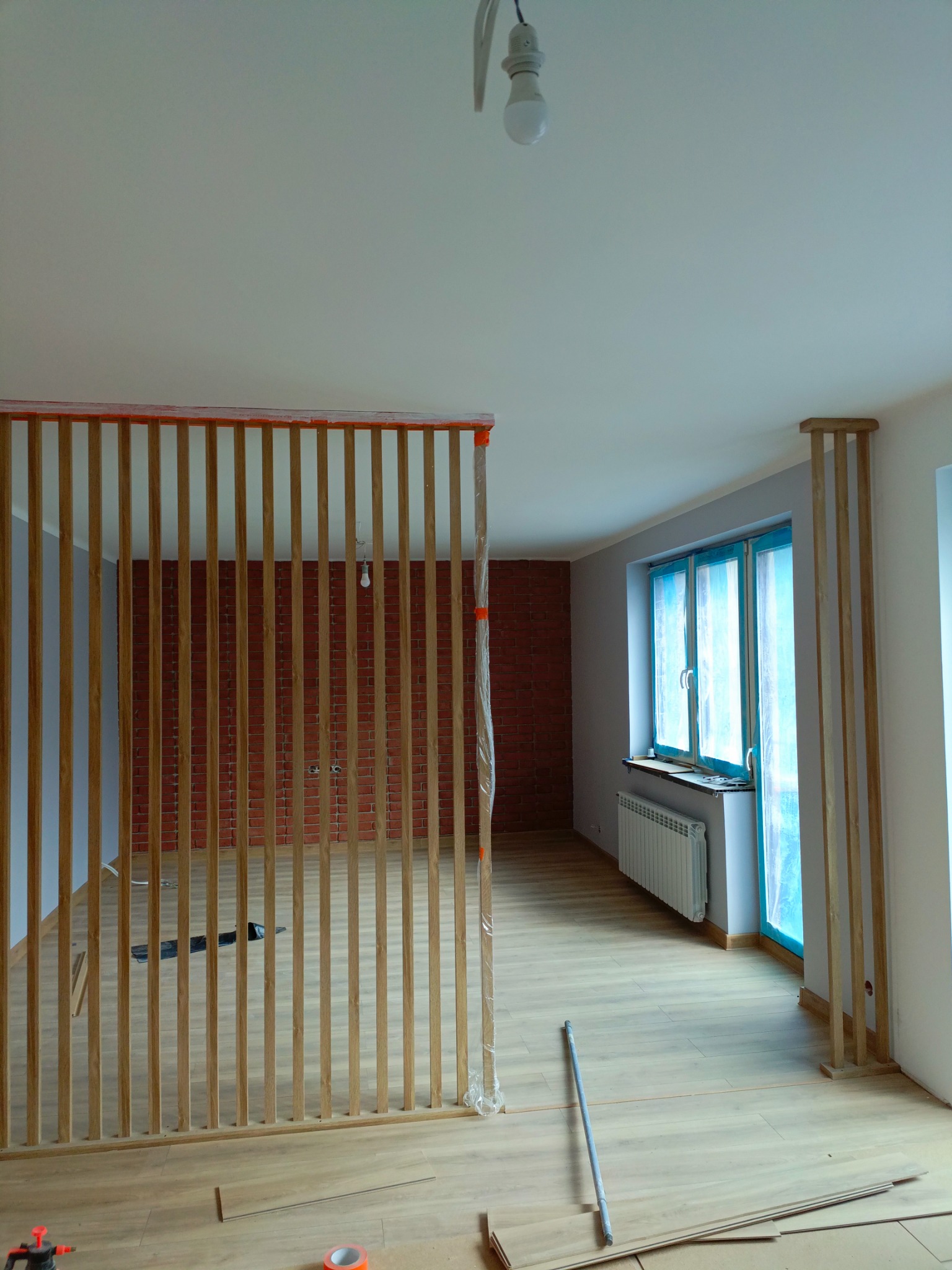 Montaż drewnianej ażurowej ścianki działowej w surowym wnętrzu z ceglaną ścianą w tle, widoczne elementy podłogi i okna zabezpieczone folią.