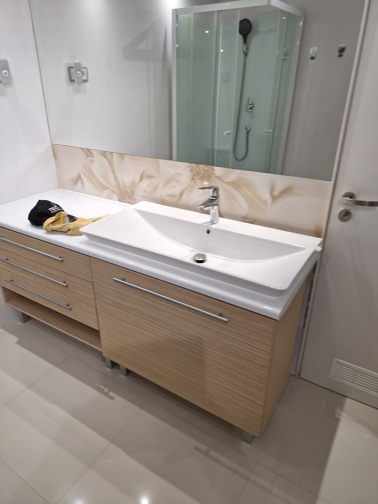 Nowoczesna łazienka z umywalką nablatową na szafce z drewnopodobnymi frontami, widoczny czarny kapelusz i żółte rękawice robocze na blacie, lustro z odbiciem kabiny prysznicowej.