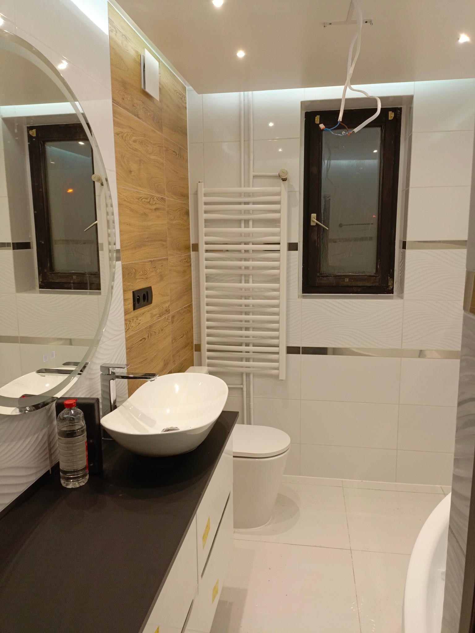 Nowoczesna łazienka z owalną umywalką nablatową, grzejnikiem drabinkowym, drewnopodobnymi płytkami na ścianie i oknem z wystającymi przewodami elektrycznymi.