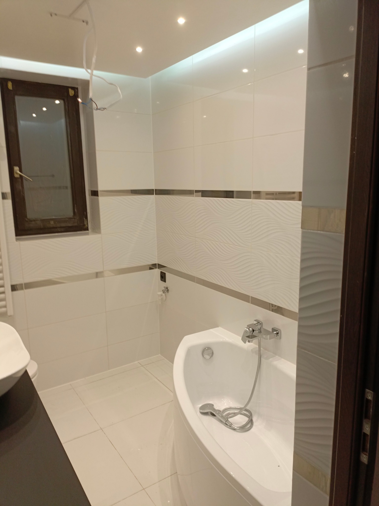 Wykończona łazienka z białą armaturą, wanną z hydromasażem, chromowaną baterią i słuchawką prysznicową leżącą w wannie. Ściany wyłożone białymi płytkami z dekoracyjnym pasem. Widoczne okno...