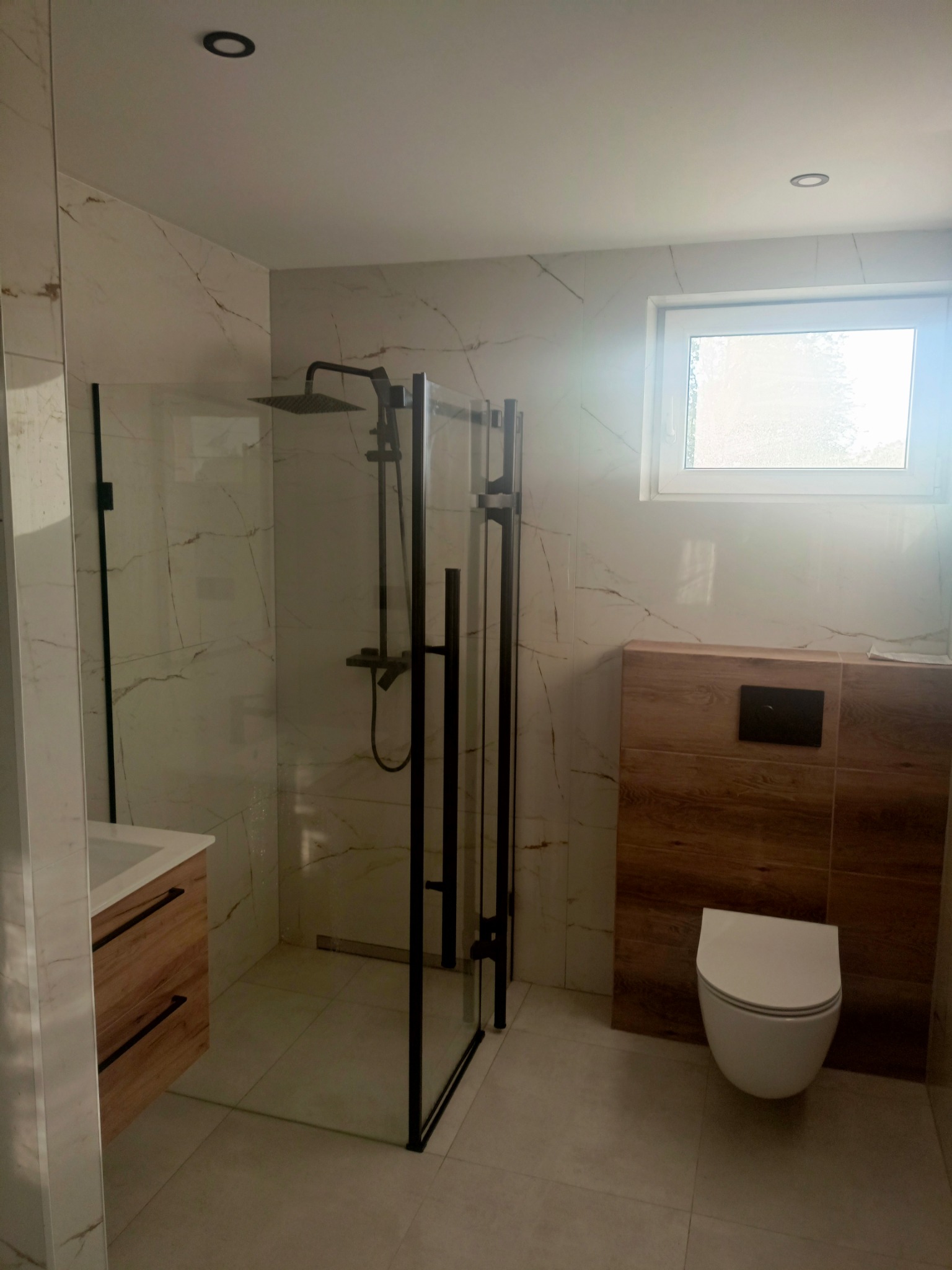 Nowoczesna łazienka z kabiną prysznicową typu walk-in z czarnymi profilami, umywalką z szafką z drewnopodobnym frontem i podwieszaną toaletą.