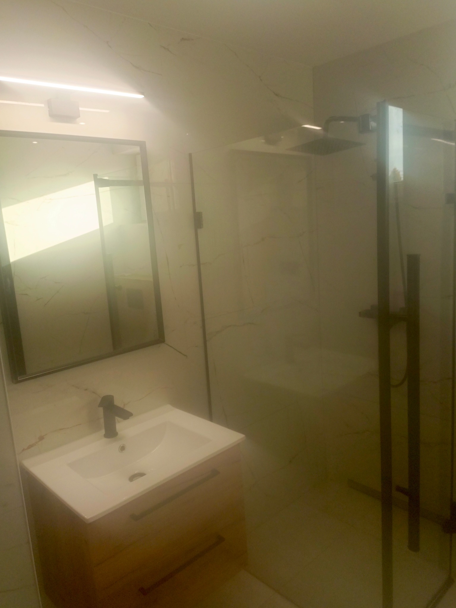 Nowoczesna łazienka z kabiną prysznicową typu walk-in, umywalką nablatową i lustrem w czarnej ramie. Marmurowe płytki na ścianach i podłodze. Minimalistyczny design.