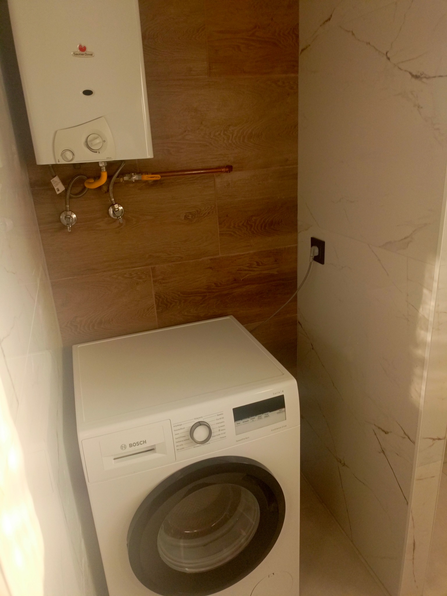 Łazienka po remoncie: biała pralka Bosch, piec gazowy Saunier Duval, brązowe i marmurowe płytki ścienne, widoczne przyłącza gazowe i wodne, gniazdko elektryczne.