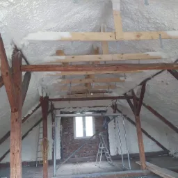 Poddasze z widoczną konstrukcją drewnianą i rusztowaniem, pokryte grubą warstwą białej pianki izolacyjnej, z fragmentem ściany z cegły i oknem.