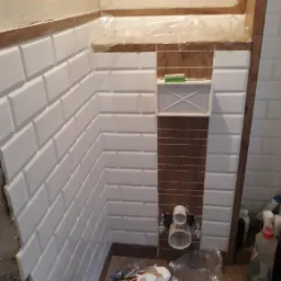 Ściany łazienki w trakcie wykańczania, z ułożonymi białymi, prostokątnymi płytkami typu metro i instalacją podtynkową WC w trakcie montażu, widoczne materiały budowlane i narzędzia.