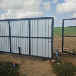 Otwarta brama przesuwna z czarną ramą i wypełnieniem z blachy trapezowej, widok na pole w tle, w pobliżu paleta i butelka wody.