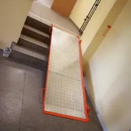 Metalowa rampa z pomarańczowym obramowaniem, ułatwiająca pokonywanie schodów, widoczna na korytarzu w budynku.