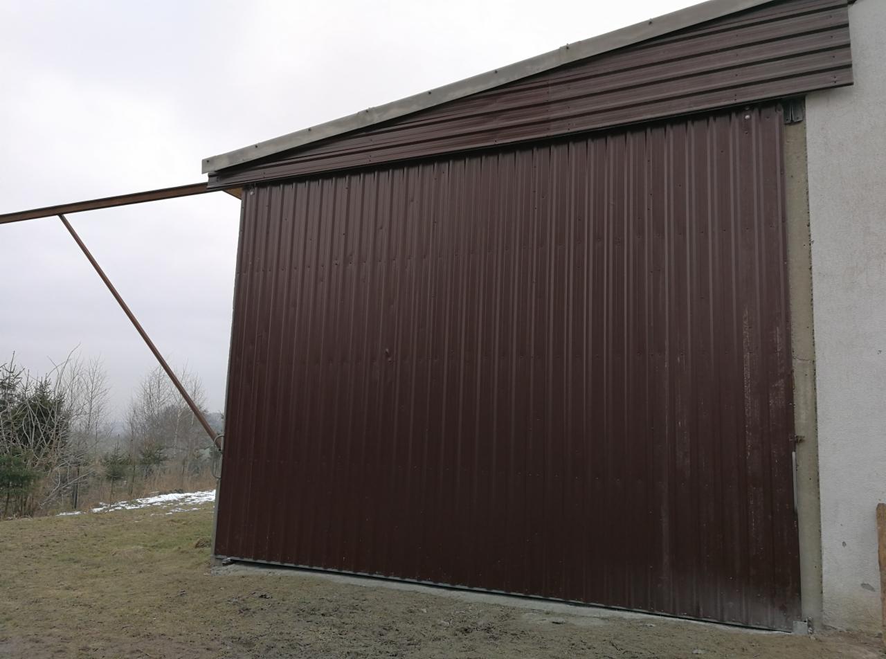 Zewnętrzna, przesuwna brama garażowa z brązowej blachy trapezowej, zamontowana przy szarym budynku z betonu, widok z zewnątrz w pochmurny dzień.
