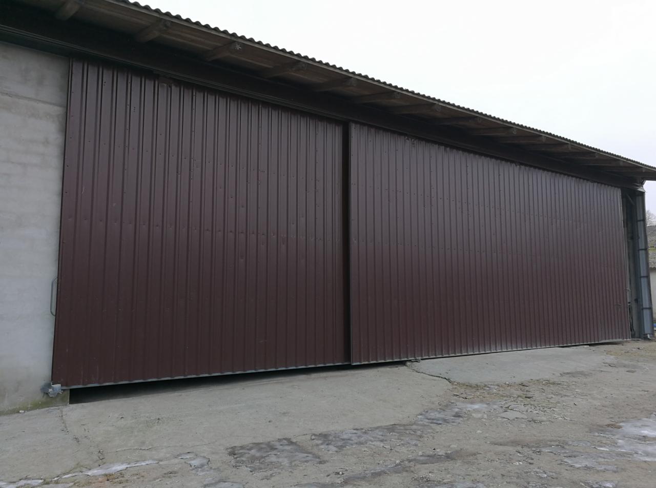 Duża, brązowa, przesuwna brama przemysłowa z blachy falistej zamontowana w szarym budynku gospodarczym, widoczna z zewnątrz na betonowym podłożu.