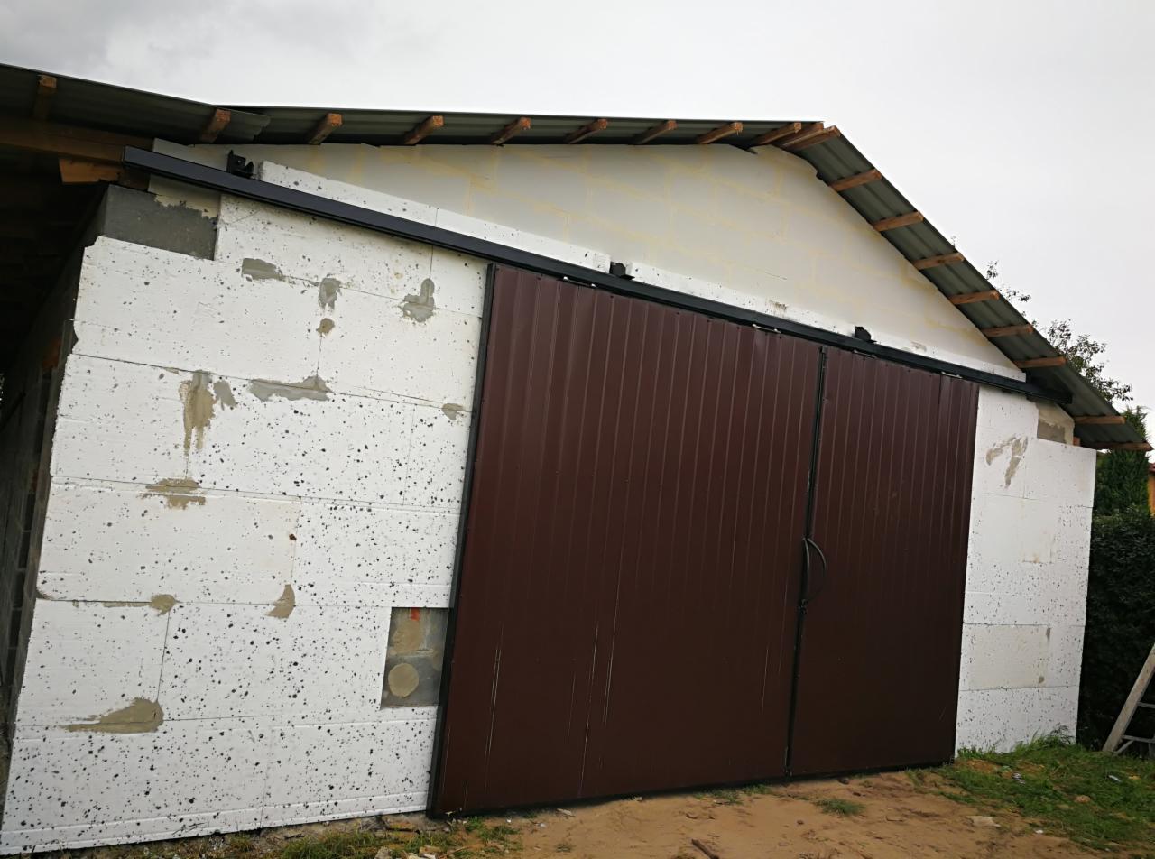 Brązowa, przesuwna brama garażowa zamontowana na budynku z białych bloczków styropianowych, widoczna czarna szyna prowadząca, teren wokół w trakcie prac wykończeniowych.