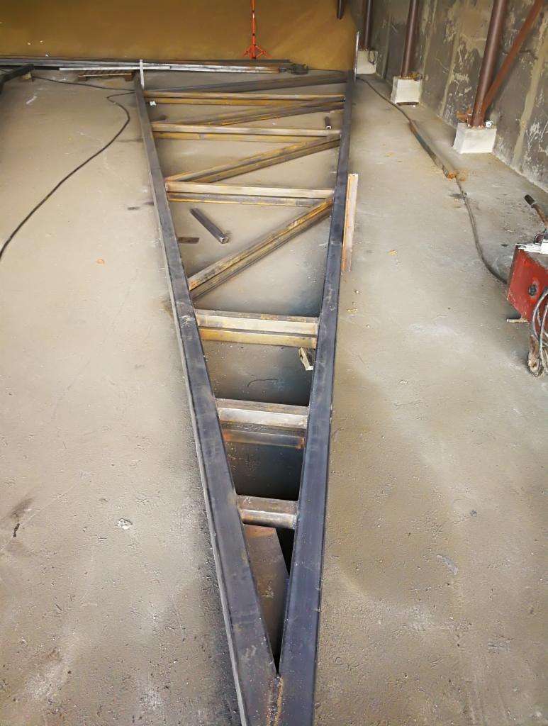 Metalowa konstrukcja kratownicowa w trakcie budowy, widoczne elementy spawane i surowy beton posadzki.