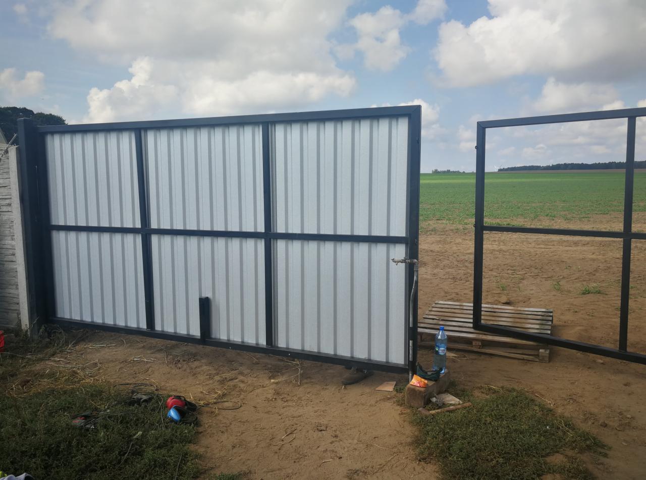 Otwarta brama przesuwna z czarną ramą i wypełnieniem z blachy trapezowej, widok na pole w tle, w pobliżu paleta i butelka wody.