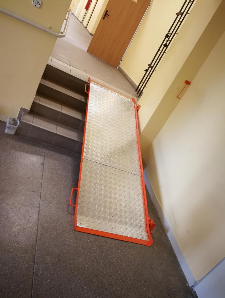 Metalowa rampa z pomarańczowym obramowaniem, ułatwiająca pokonywanie schodów, widoczna na korytarzu w budynku.