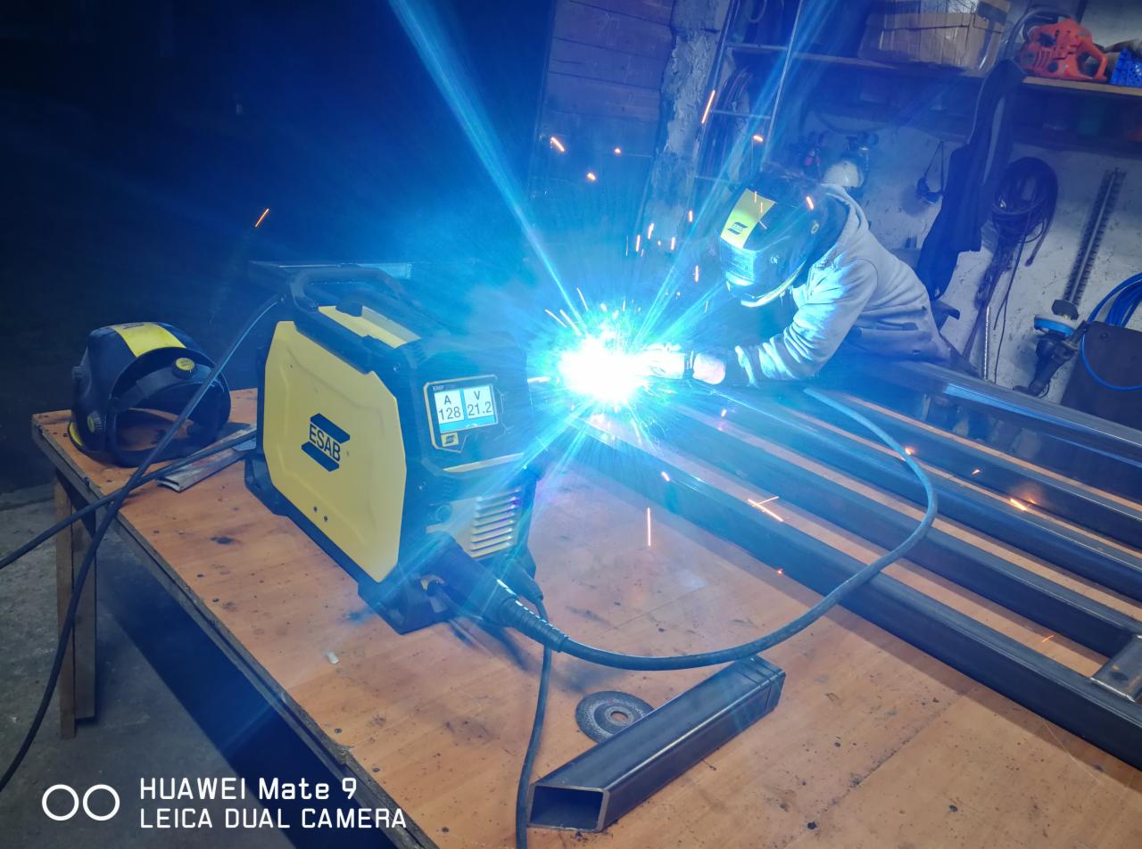 Pracownik w masce spawalniczej łączy metalowe profile za pomocą urządzenia spawalniczego ESAB, iskry i intensywne światło podczas spawania, na stole roboczym leży dodatkowa maska i fragment profilu.