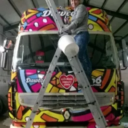 Mężczyzna na drabinie maluje kolorową grafikę na przodzie samochodu dostawczego w warsztacie.