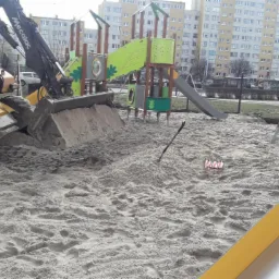 Żółta koparko-ładowarka Mecalac pracuje na placu budowy placu zabaw, wyrównując teren pokryty piaskiem, w tle blok mieszkalny i elementy konstrukcji placu zabaw.