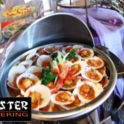 Bufet z podgrzewaczem, danie z przegrzebków w muszlach, udekorowane brokułami i papryczką chili, na stole z fioletową dekoracją.