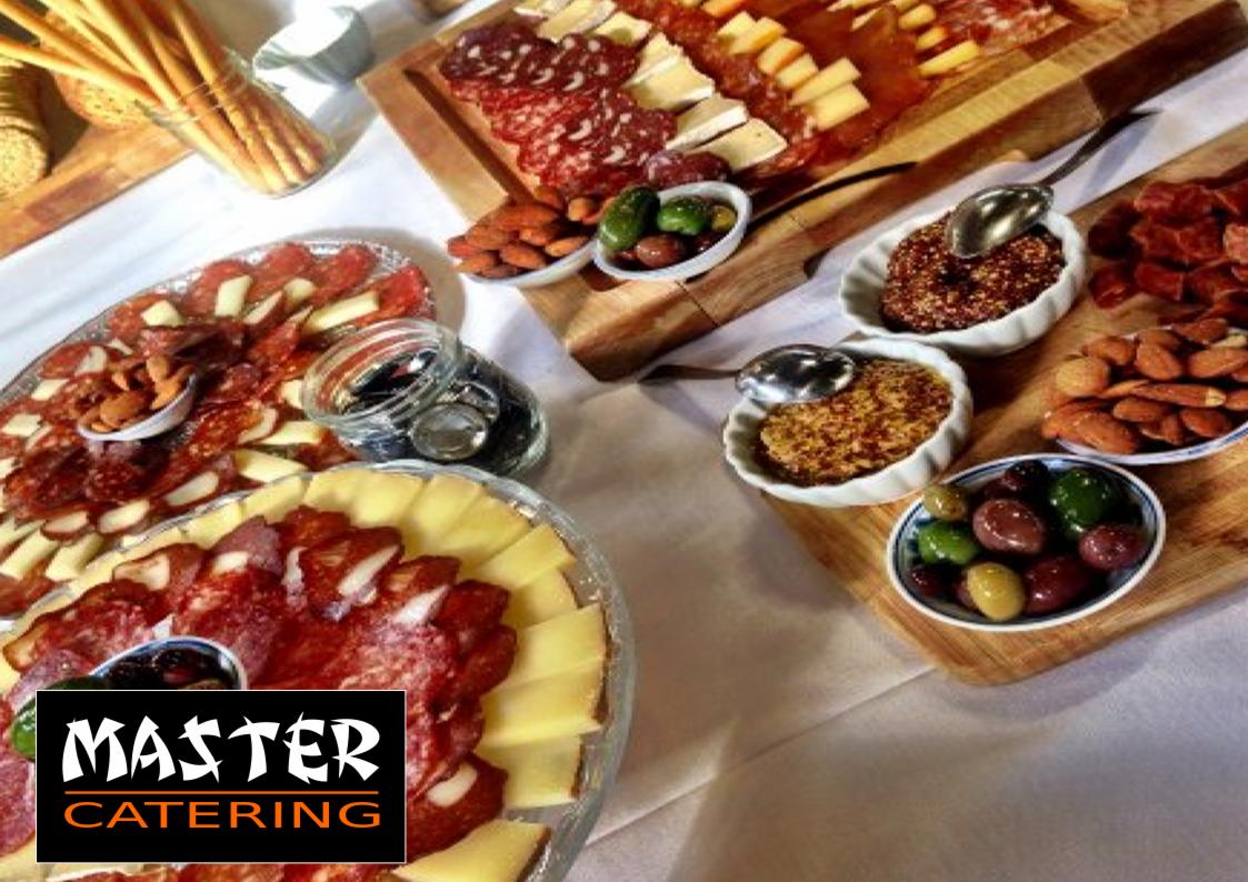 Bogato zastawiony stół z deskami serów, wędlin, oliwek i migdałów, z widocznym logo firmy cateringowej w lewym dolnym rogu.
