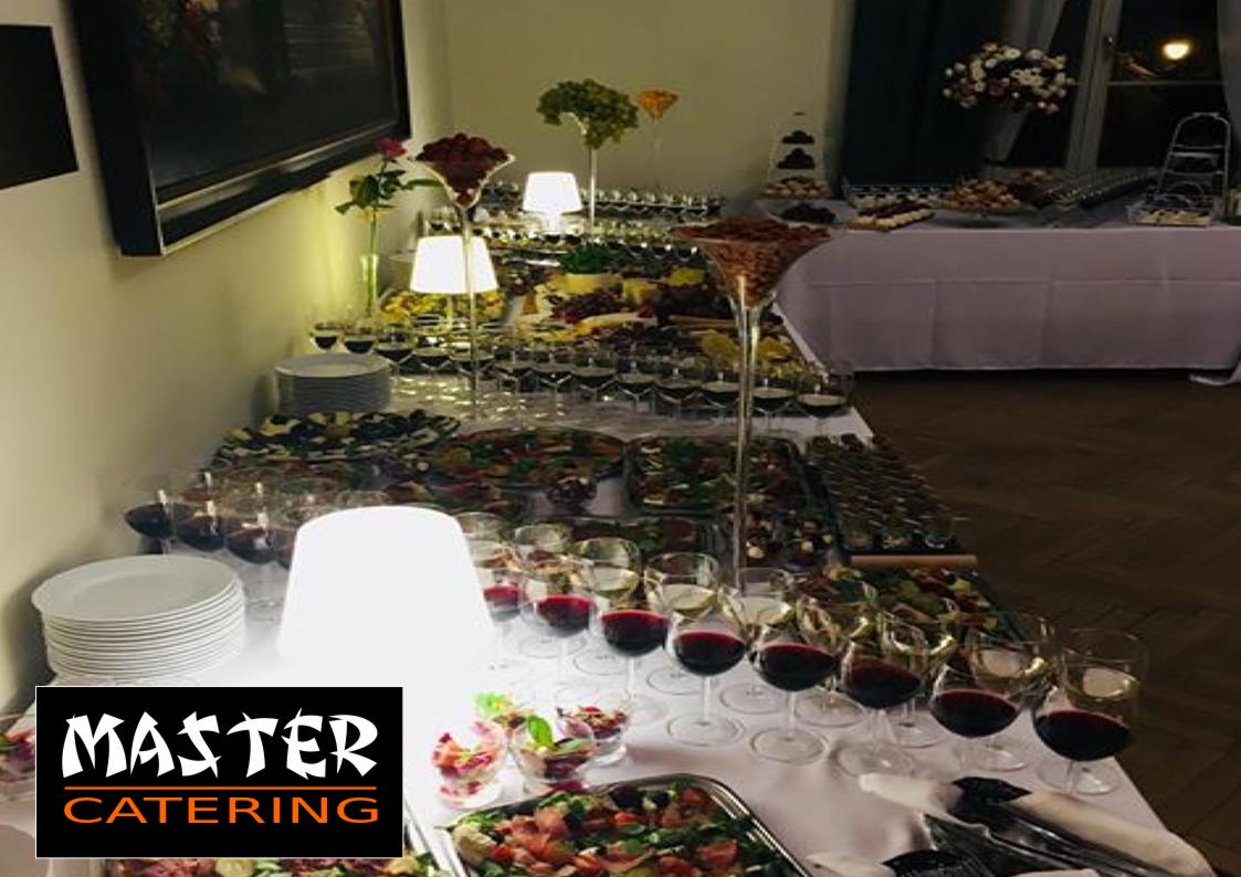 Bogato zastawiony stół z różnorodnymi przekąskami, deserami, owocami i napojami, w tym kieliszki z czerwonym winem, podświetlony lampami, z widocznym logo firmy cateringowej.