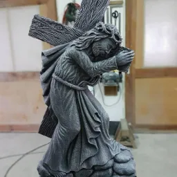 Szara kamienna rzeźba Jezusa niosącego krzyż, z widoczną fakturą drewna na krzyżu i drapowaną szatą, umieszczona na podstawie imitującej skały, w tle warsztat kamieniarski.