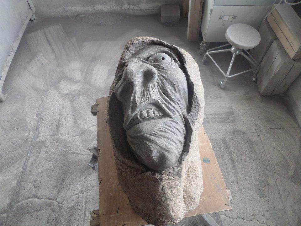 Rzeźba kamienna przedstawiająca ekspresyjną twarz z otwartymi ustami i wyrazistymi zmarszczkami, umieszczona w surowym, warsztatowym otoczeniu z betonową podłogą i widocznym stołem roboczym.