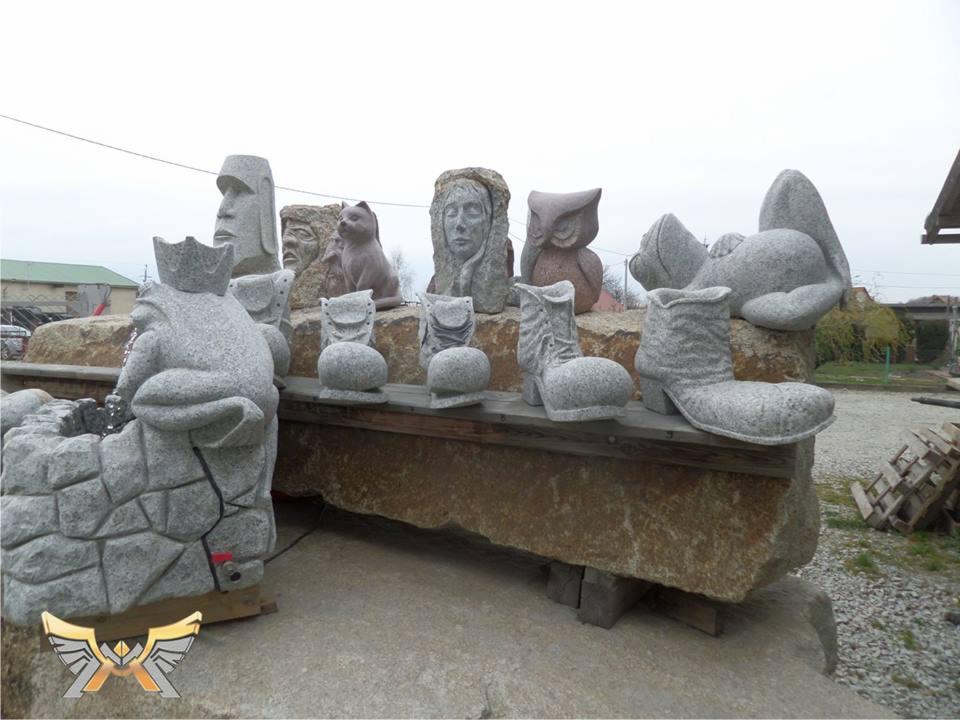 Różnorodne rzeźby z szarego kamienia, w tym głowy, zwierzęta (kot, sowa, królik) i buty na obcasie, ustawione na kamiennej ławie, z fontanną z żabą w koronie po lewej stronie.