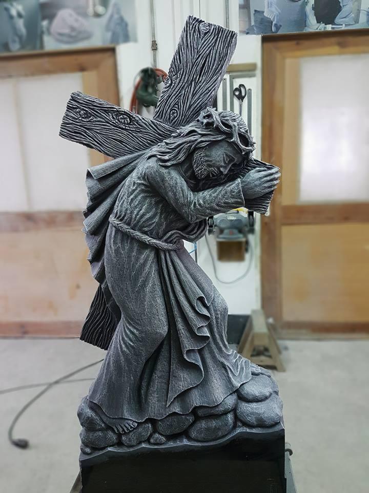 Szara kamienna rzeźba Jezusa niosącego krzyż, z widoczną fakturą drewna na krzyżu i drapowaną szatą, umieszczona na podstawie imitującej skały, w tle warsztat kamieniarski.