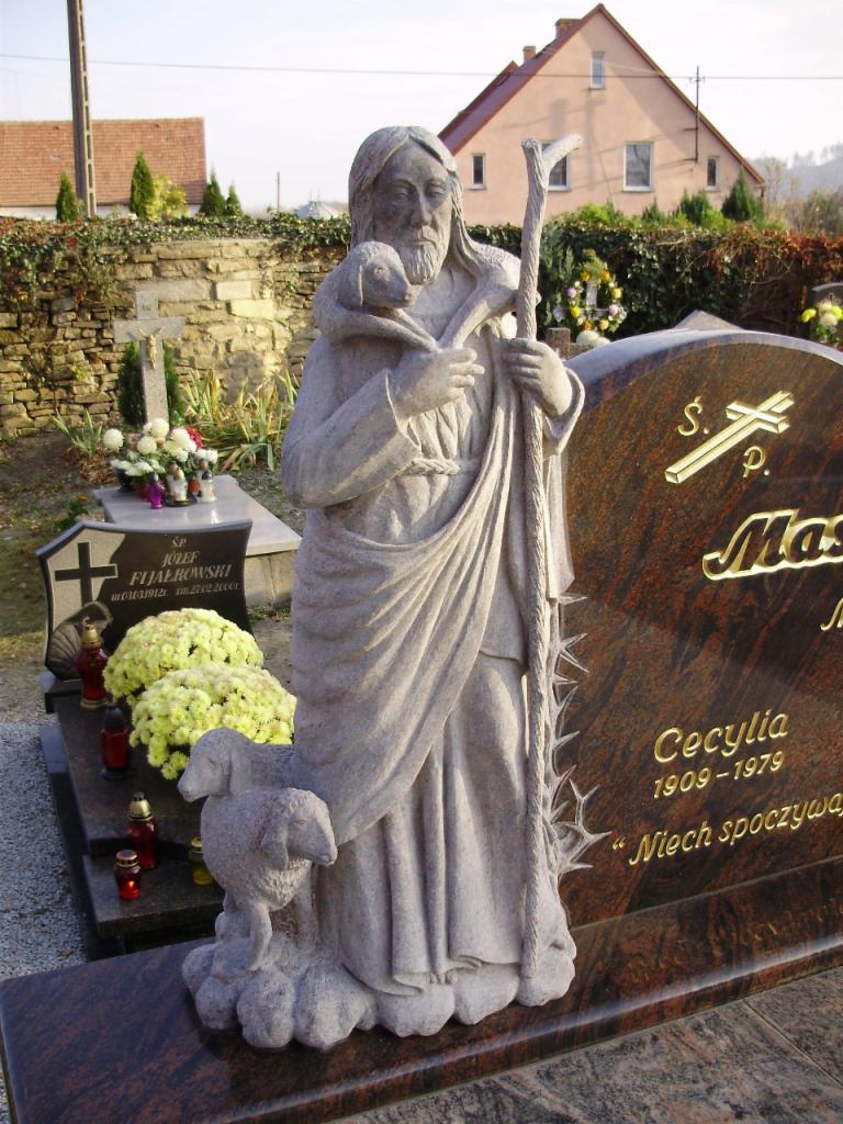 Granitowy pomnik z rzeźbą Jezusa Dobrego Pasterza z owieczką na ramionach, ustawiony na ciemnym, polerowanym nagrobku z inskrypcjami.