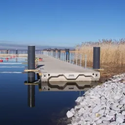Nowoczesny, betonowy pomost pływający z czarnymi słupami, częściowo zanurzony w wodzie, z widocznymi elementami konstrukcyjnymi i odbiciem w tafli wody, otoczony kamieniami i trzciną w tle.