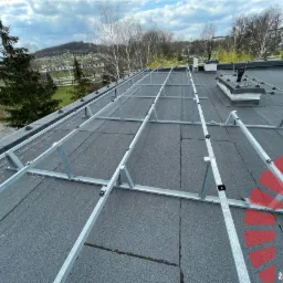 Montaż konstrukcji wsporczej z powłoką Magnelis® dla paneli bifacialnych na dachu pokrytym papą, widok z góry na tle zabudowań miejskich i drzew.