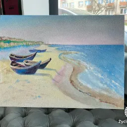 Obraz przedstawiający plażę z łodziami, namalowany w pastelowych odcieniach, umieszczony na szarym, pikowanym fotelu. W tle widoczne okno z budynkiem i samochodem.