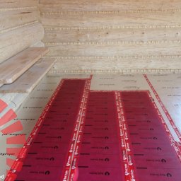 Życie na prąd sp. z o.o. - Montaż folii grzewczych Red Snake na podłodze w drewnianym domu z bali, widoczne panele podłogowe przygotowane do montażu.