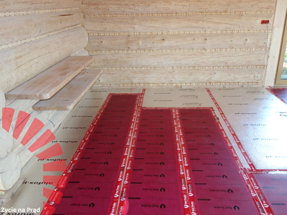 Montaż folii grzewczych Red Snake na podłodze w drewnianym domu z bali, widoczne panele podłogowe przygotowane do montażu.