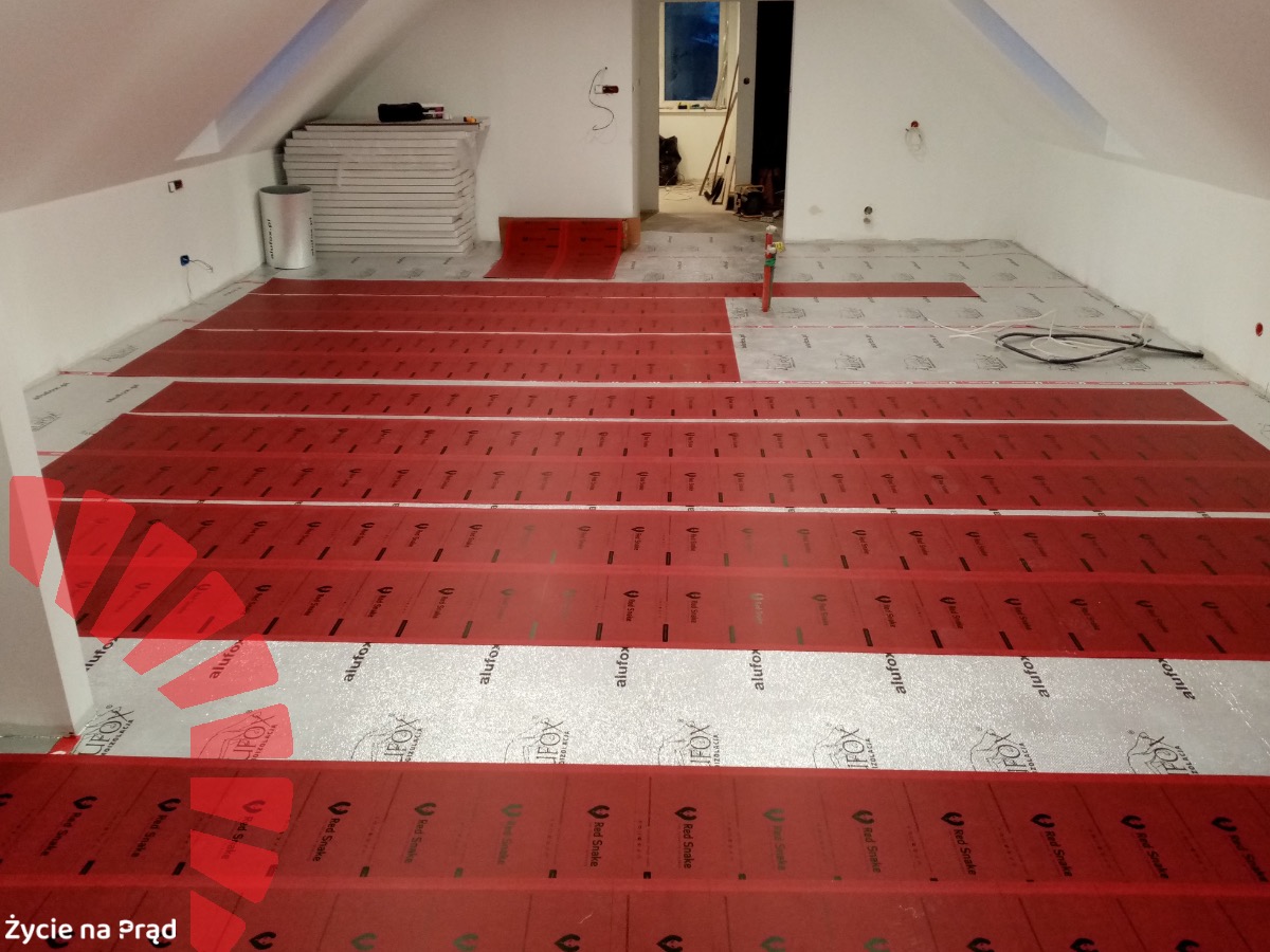 Montaż folii grzewczej na podczerwień na podłodze poddasza, widoczne rozłożone arkusze folii z czerwonymi paskami i srebrną izolacją, przygotowane do dalszych prac instalacyjnych, z widocznymi...