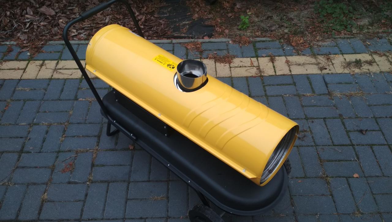 Żółta nagrzewnica powietrza na czarnej podstawie z kółkami, stojąca na szarej kostce brukowej na zewnątrz.