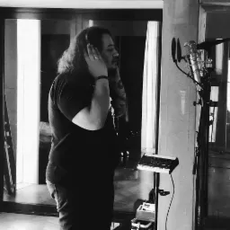 Czarno-białe zdjęcie wokalisty w słuchawkach, stojącego przed mikrofonem w pomieszczeniu z akustycznymi panelami.