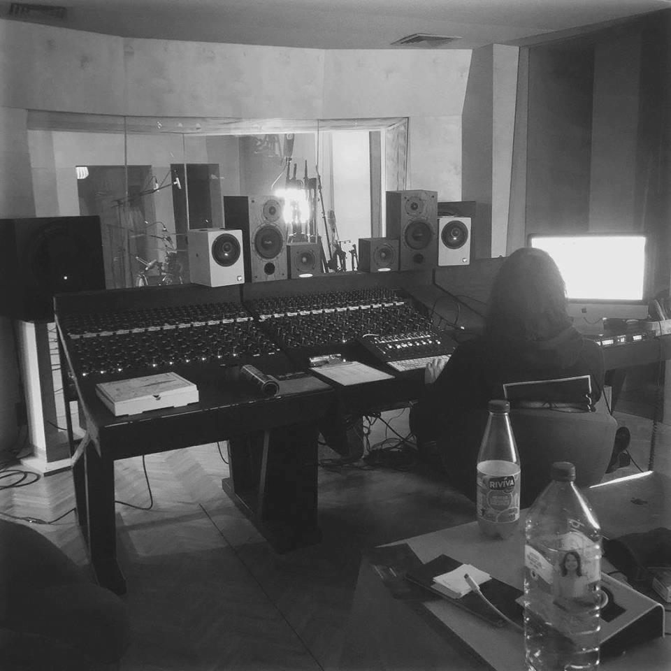 Czarno-białe zdjęcie wnętrza pomieszczenia z dużą konsolą mikserską, licznymi głośnikami ustawionymi na półkach za konsolą oraz postacią siedzącą przed ekranem komputera; na pierwszym planie stolik...