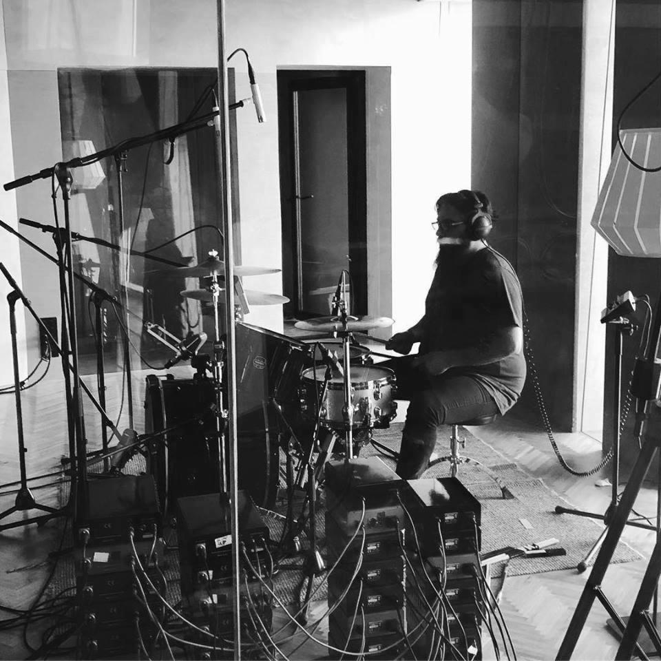 Czarno-białe zdjęcie perkusisty w słuchawkach, grającego na perkusji w pomieszczeniu z panelami akustycznymi i mikrofonami, widoczne kable i sprzęt audio.