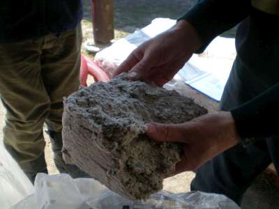 Bryła szarej wełny mineralnej trzymana w dłoniach, widoczna struktura warstw, w tle osoba w roboczych spodniach, Bochnia.