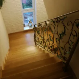 Drewniane schody z ozdobną, metalową balustradą z motywami roślinnymi, widok z góry, okno z dekoracjami w tle.