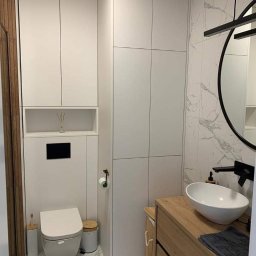 Mateusz Trepczyk Stol-Trep - Nowoczesna łazienka z zabudową meblową: wysoka biała szafa z półką, podwieszana toaleta, drewniana szafka z umywalką nablatową i marmurowe płytki.