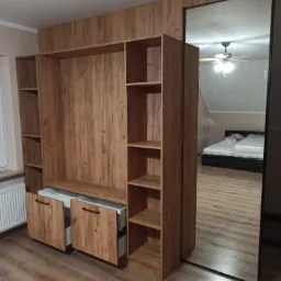 Drewniana szafa wnękowa z lustrem odbijającym sypialnię z łóżkiem i wentylatorem sufitowym, częściowo otwarte szuflady.