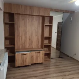 Zabudowa meblowa z drewnopodobnymi frontami, z otwartą szufladą i półkami, z lustrem po prawej stronie, widok na wnętrze pokoju przez lustro.