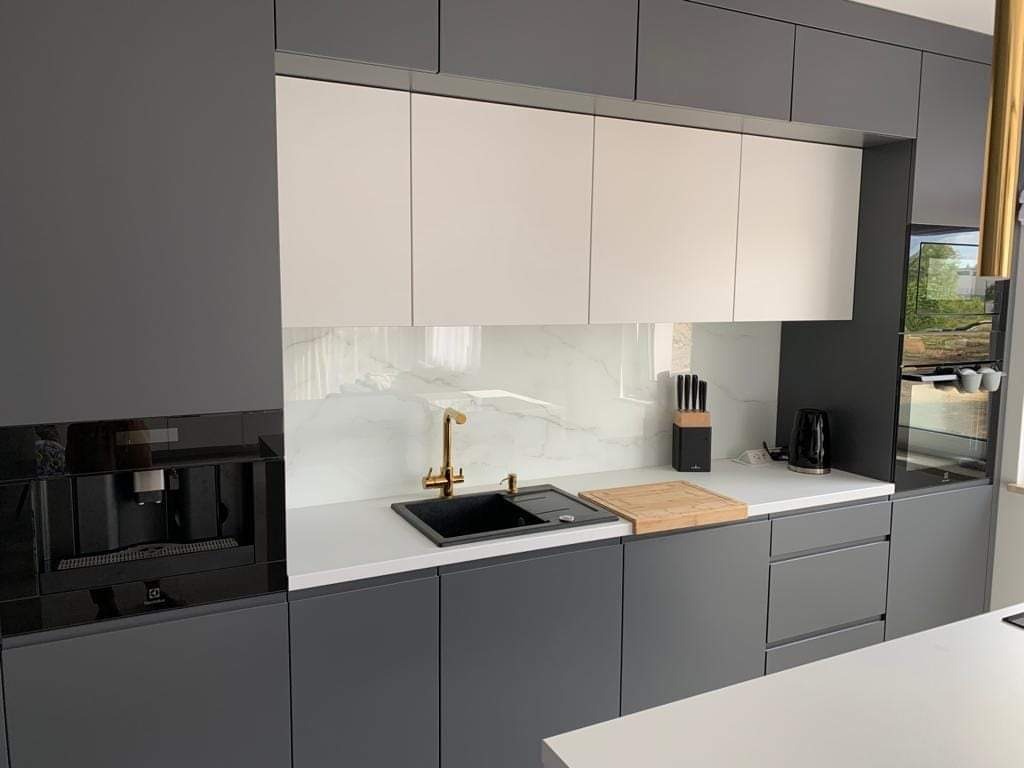 Nowoczesna szara kuchnia z białymi szafkami górnymi, marmurowym backsplash, złotą baterią i wbudowanym ekspresem do kawy.